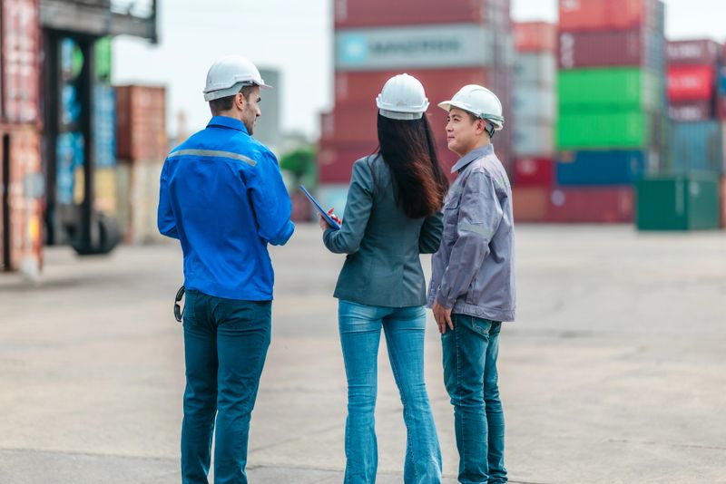 The female manager supervises and discusses logistics with two male workers at shipping container yard. Stacked containers and industrial equipment, highlight global trade and transportation industry