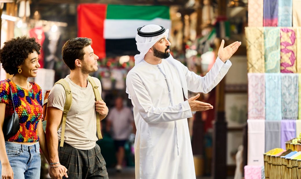 A man in traditional Emirati dress shows textiles to tourists in a market with UAE flag.