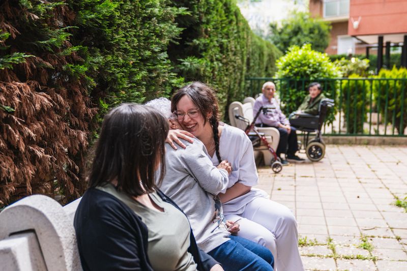 Nurse is hugging an elderly woman on a bench in the garden of a nursing home, providing comfort and care