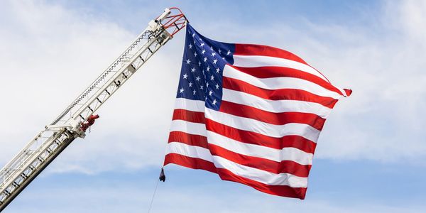 Large American flag flying from a fire truck ladder against a blue sky.