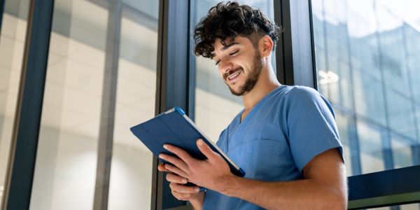 A smiling healthcare worker using a tablet near large windows.