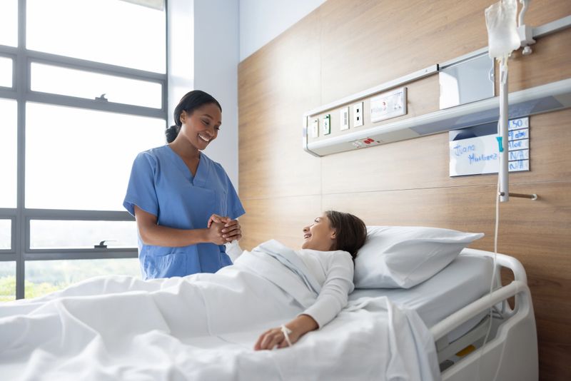 Friendly African American nurse holding the hand of girl lying in bed at the hospital - healthcare and medicine concepts