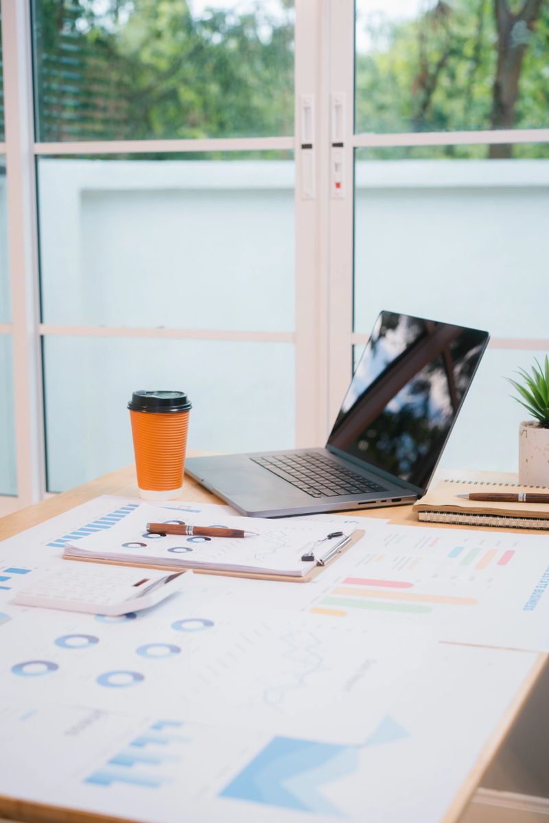 A bright, modern office space with a wooden desk covered in documents, charts, a laptop, and a coffee cup. A white chair and large windows create a clean, productive atmosphere.