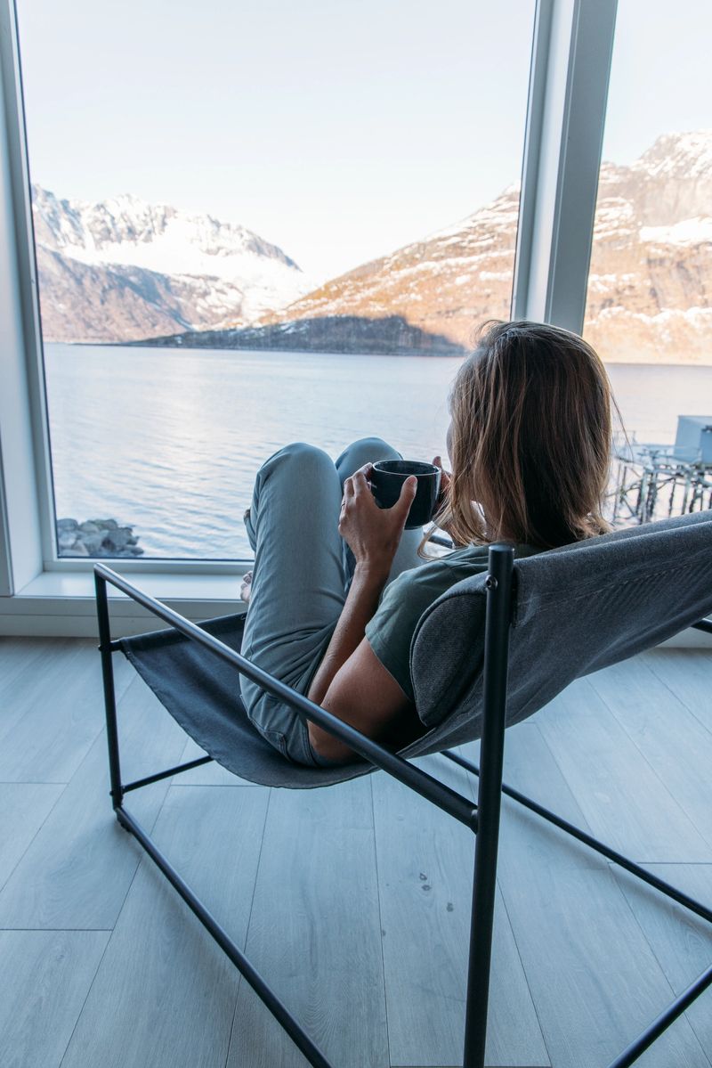 Woman enjoying a warm drink by a large window with a view of a serene Scandinavian fjord in springtime. Calming waters and snow-capped mountains. Peaceful retreat in Northern Europe.