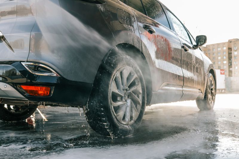 Modern SUV being cleaned with high-pressure water and foam at a self-service car wash, rear side view.