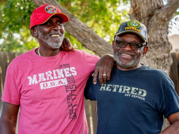 Two smiling veterans wearing military-themed shirts and hats outdoors.