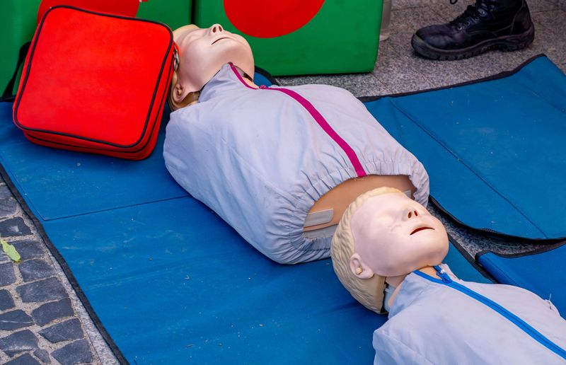 Two realistic CPR training dummies lying on blue exercise mats, set up for a first aid demonstration or resuscitation course. Visible emergency bag and instructor’s shoes. Educational setup for teaching life-saving techniques in public or outdoor setting.
