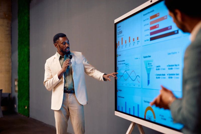 Businessman is holding a microphone and pointing at a large interactive display showing sales data during a conference, while another businessman watches