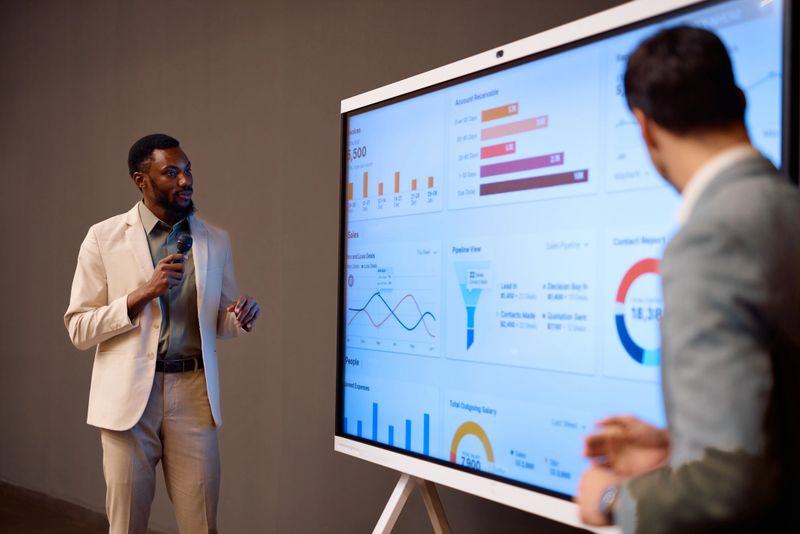 Businessman presenting company performance data on large interactive display during a meeting with colleagues, using a microphone and showing graphs, charts and key performance indicators