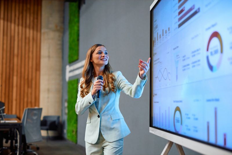 Businesswoman is holding microphone and pointing at interactive whiteboard, presenting marketing data during a business meeting in the office