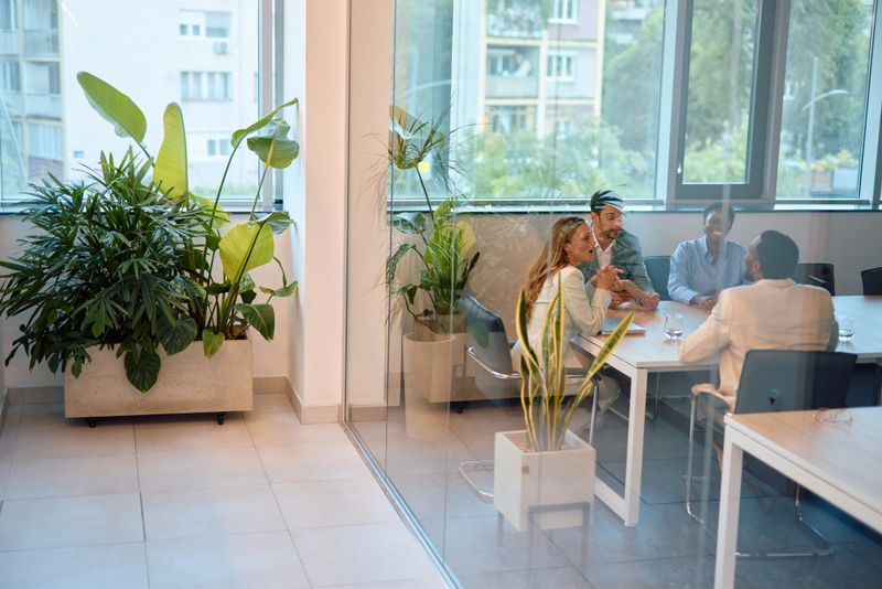 Business team engaged in lively discussion during a meeting in a modern office, surrounded by plants, sitting around a glass table, sharing ideas and collaborating effectively