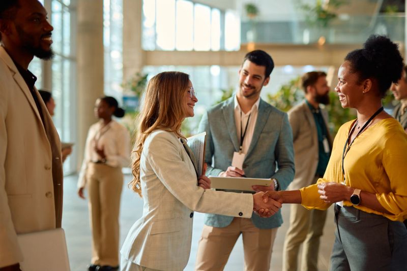 Two businesswomen shaking hands during a conference meeting, engaging in networking while other professionals converse in the background, highlighting collaboration and partnership