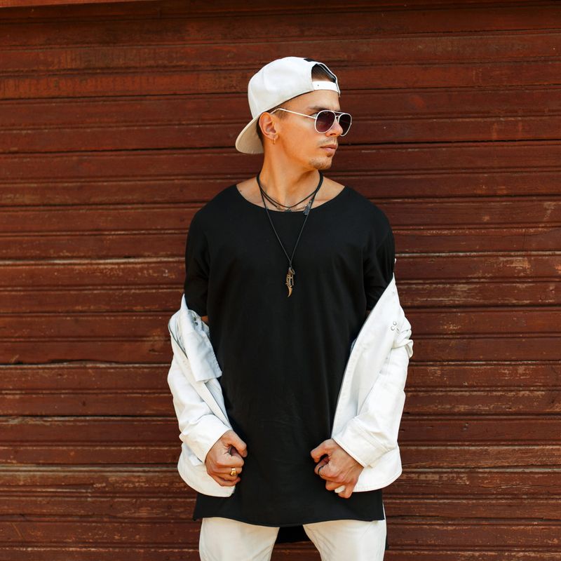 Fashionable handsome man in a black T-shirt with a white baseball cap and sunglasses posing near a wooden wall