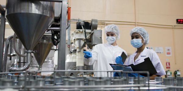 Dos mujeres en bata y mascarilla inspeccionan maquinaria industrial.