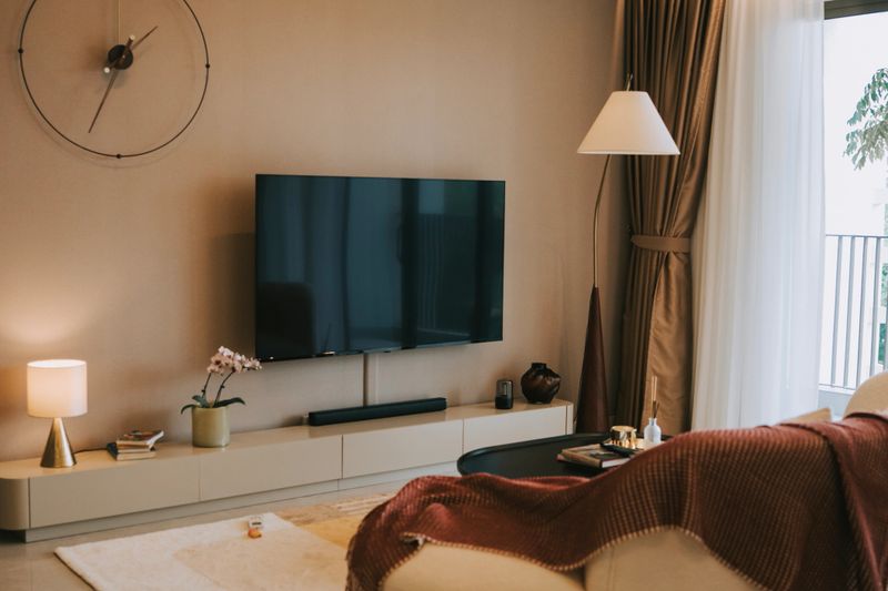 Stylish and minimalistic living room interior featuring a cream-colored curved sofa with neutral cushions and a brown throw blanket. The space is complemented by a round black coffee table, floor-to-ceiling glass doors, and soft natural lighting. Elegant decor includes a large round mirror, indoor plants, and a serene balcony view, creating a warm and inviting atmosphere.