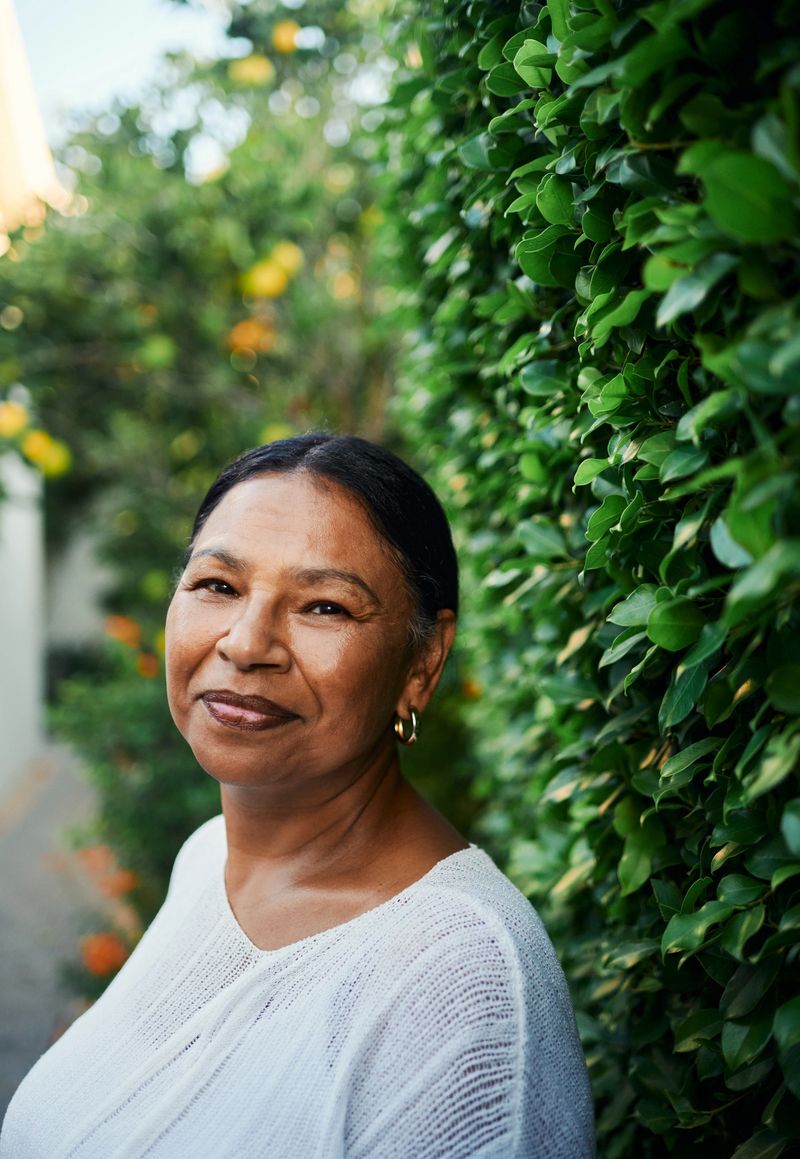 A middle-aged woman with warm expression stands in a vibrant green garden. She wears a white top and gold earrings, surrounded by nature.