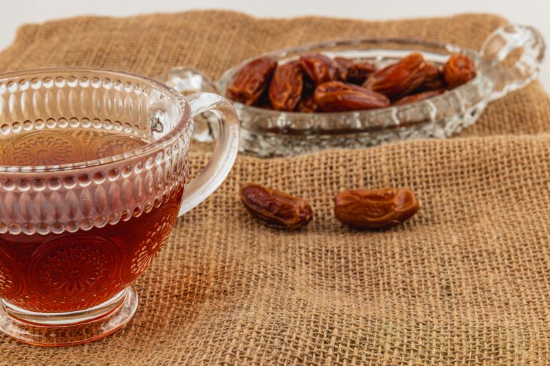 A clear glass cup filled with dark tea sits next to a delicate dish of dates on a textured burlap cloth. The setting evokes a cozy atmosphere ideal for relaxation.