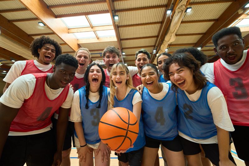 Multi-ethnic high school basketball team members shouting, holding basketball, and making eye contact in gym