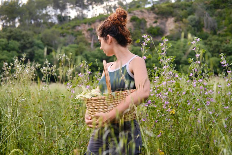 Young woman walking through a field of flowers, carrying a wicker basket full of freshly picked herbs