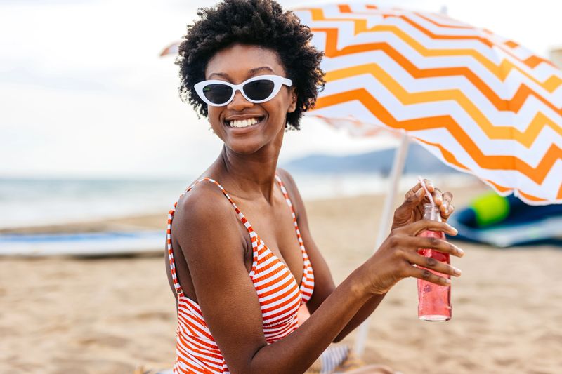 Portrait of a young black woman drinking a fizzy drink while relaxing underneath a parasol on the beach in Spain.
