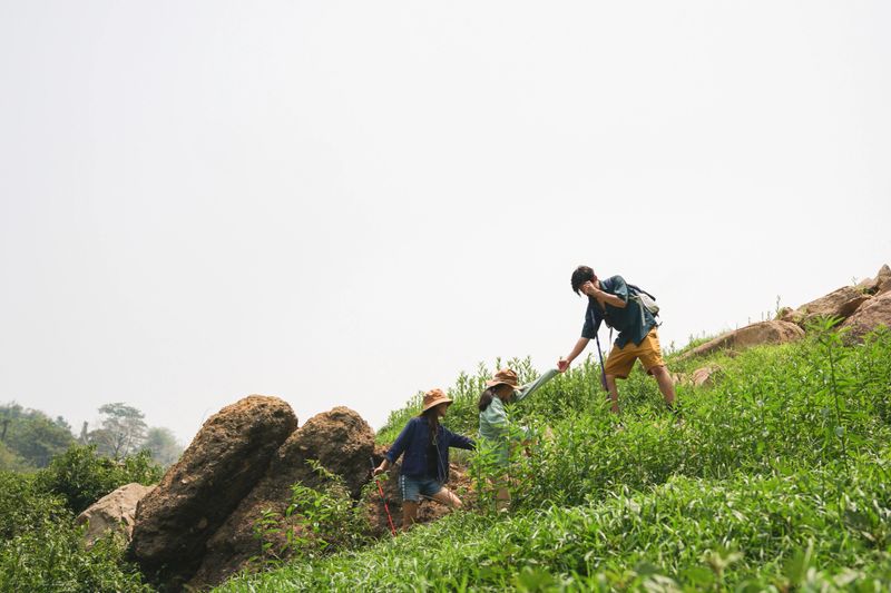 Group of Friends Adventure Hiking on Mountain Trail in Summer, Man Helping Each Other While Trekking Outdoors, Outdoor Teamwork and Exploration on a Green Hillside