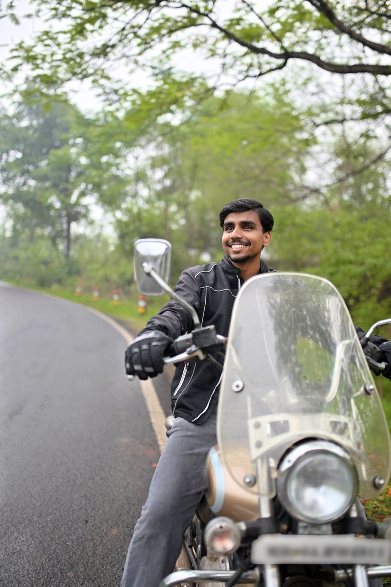 Indian man in a black riding jacket seated on a classic motorcycle, looking off into the distance with a smiling and confident expression. Set against a lush green backdrop on a rural road with another biker passing in the background, the scene evokes a sense of freedom, tranquility, and the open road.