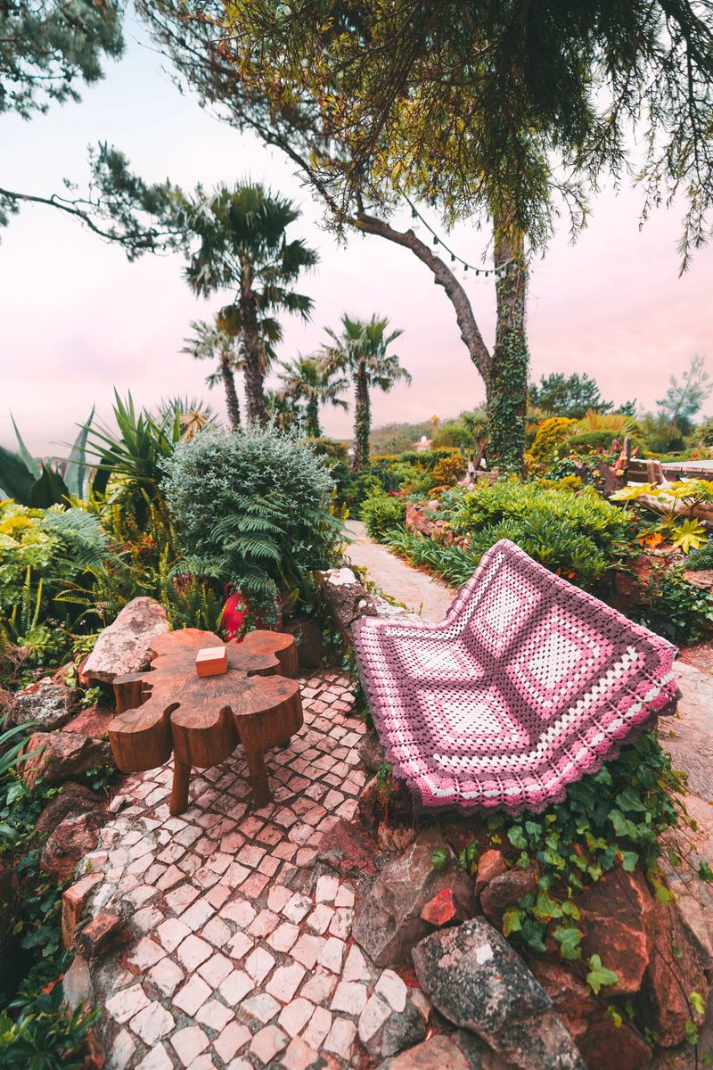 Vertical wide-angle view of a colorful garden nook with a handmade crochet blanket draped over a rustic stone bench, a flower-shaped wooden table, lush tropical greenery, and palm trees at sunset