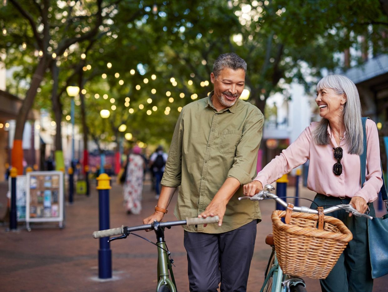 Older couple smiling and walking with bicycles in a tree-lined street.