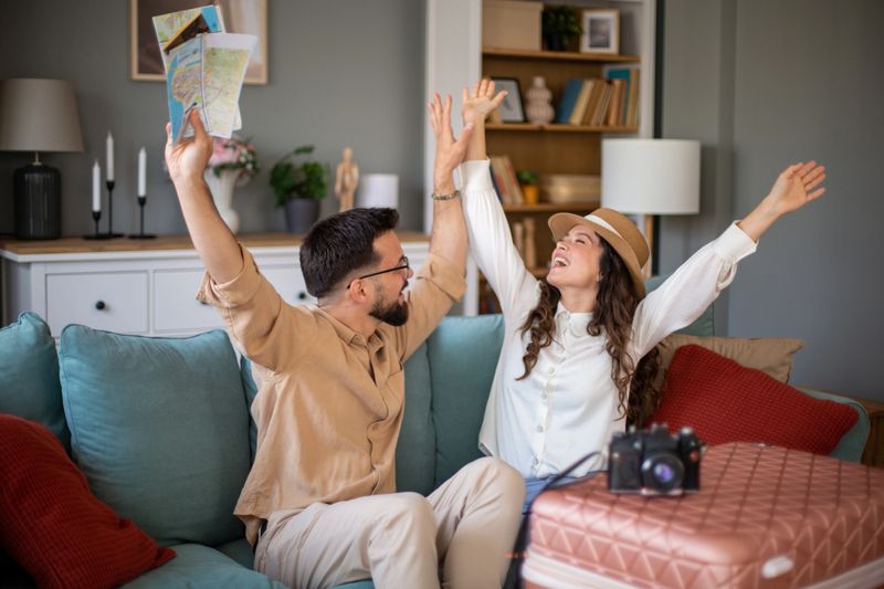 Young couple sitting on a sofa and holding hands, celebrating upcoming trip, holding maps and raising arms