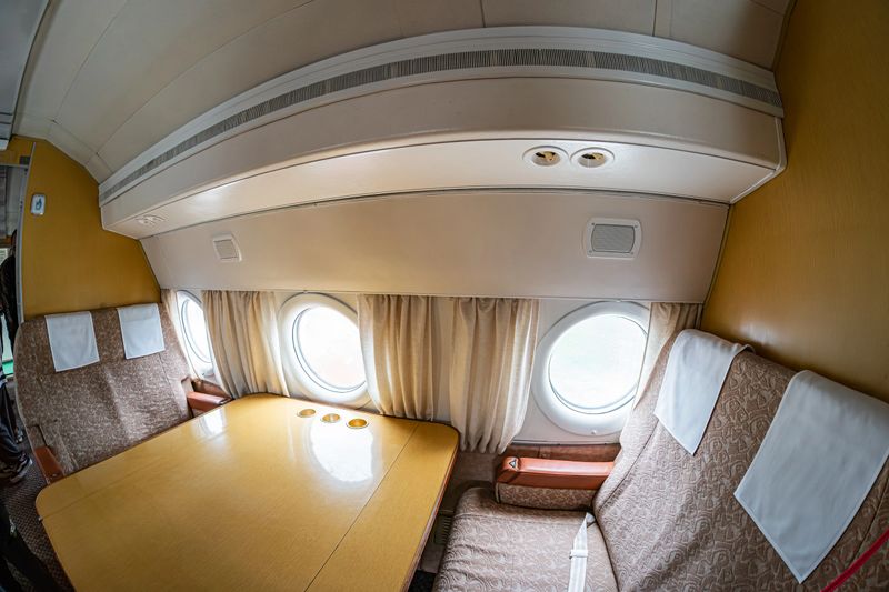 Vintage interior of a small airplane cabin with light colored walls, metal ductwork, partially drawn curtains at a window, multiple rows of white and brown seats, carpeted floor, and a central table