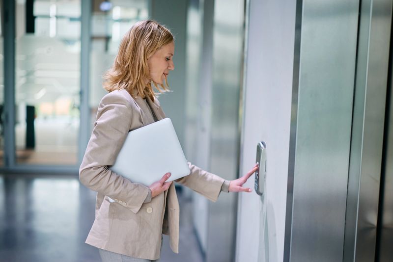 Businesswoman is calling the elevator in an office building, she's pressing the button while holding a laptop under her arm, waiting for the elevator to arrive