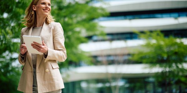 Smiling woman in a beige suit holding a tablet outdoors near modern architecture.