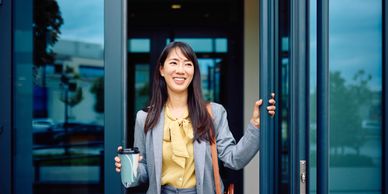 A smiling woman in business attire holding a coffee cup while opening a glass door.