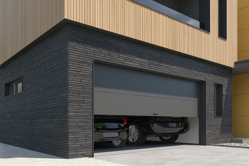 Front-facing view of a contemporary double garage, designed for two vehicles, featuring dark grey to black sectional roll-up doors. The facade is accented with sleek black slate stone, adding texture and contrast to the clean, modern lines of the home. The entrance design is minimalist and functional, blending seamlessly with the overall architectural style. Ideal image for use in showcasing modern garage solutions, facade design, urban housing concepts, or high-end real estate and construction materials.