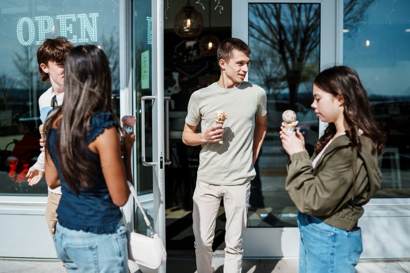 A group of teenage boys and girls exit an ice cream shop onto a sunny sidewalk while holding ice cream cones.