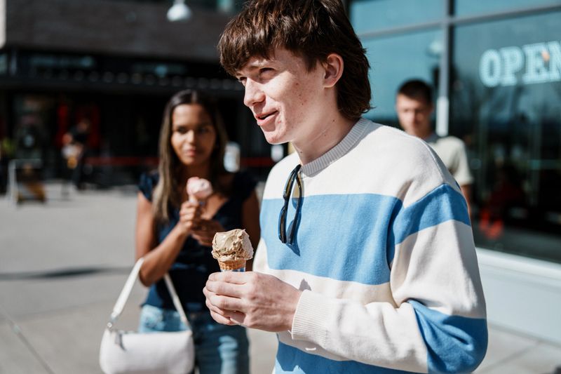 A Caucasian teenage boy chats with his friends as they eat ice cream cones and hang out on the sidewalk in front of the shop on a sunny afternoon.