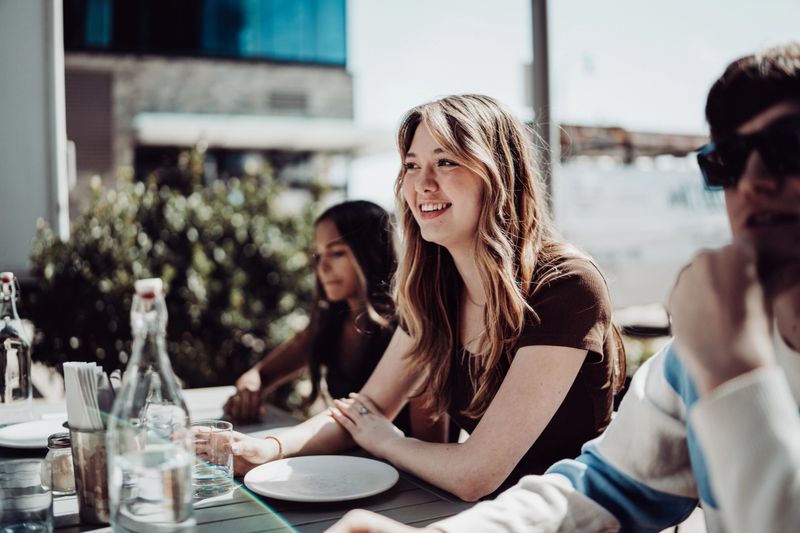 A Caucasian teenage girl who is dining outdoors at a pizzeria with friends, smiles as the group chats and waits for their food to be served.
