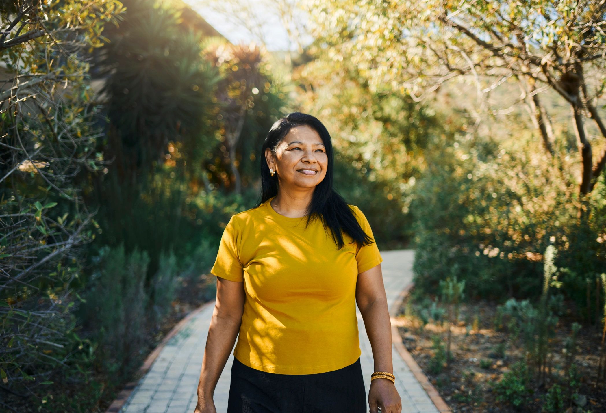Mujer sonriente con camiseta amarilla en un camino al aire libre.