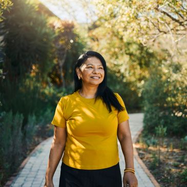 Smiling woman in yellow shirt walking on a sunny garden path.