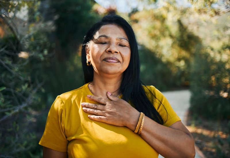 A serene middle-aged woman with dark hair and diverse features wears a yellow shirt, meditating peacefully in a lush outdoor environment under soft sunlight.
