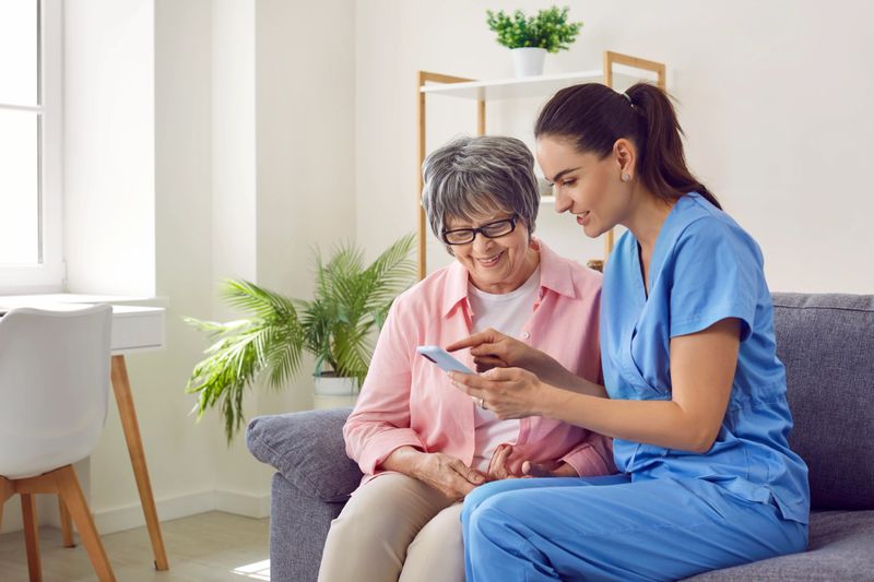 Young pretty nurse at home shows an elderly woman a photo on a mobile phone and tells positive happy moments of life, conveying a good mood and a positive attitude. Concept of psychological health.