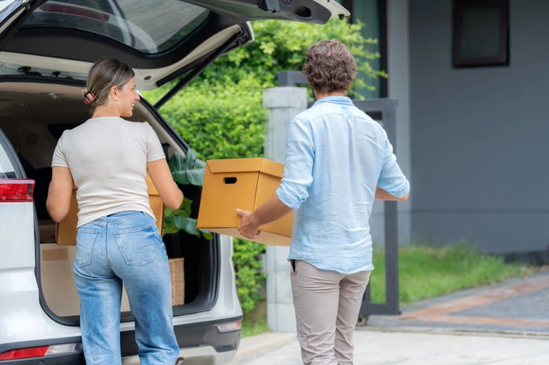 Smiling young couple packs belongings into a car, preparing to move into a new house. Man holds cardboard box while woman helps organize. A joyful relocation moment symbolizing love, beginnings, support, and life transitions.