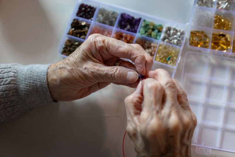Elevated close-up view of a senior female's hands, which appear old and delicate as she carefully tries to thread a bead, attempting to create a beaded piece of jewellery with a selection of multi-coloured beads in a partitioned, boxed container in front of her.