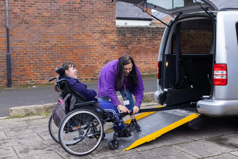 A happy young woman sitting in her wheelchair waiting as her female friend, a care assistant who has brought her to a community centre for the day is preparing the mobility car and wheelchair in the carpark for the journey home. The young woman has a condition called Quadriplegic cerebral palsy that affects all four limbs. She also has moderate learning disability.