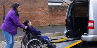 Woman helps man in wheelchair onto a van ramp.