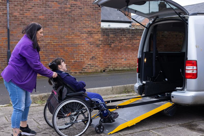 A happy young woman sitting in her wheelchair waiting as her female friend, a care assistant who has brought her to a community centre for the day . The assistant carer is pushing the wheelchair up the ramp into the rear of the mobility vehicle in a carpark for the journey home. The young woman has a condition called Quadriplegic cerebral palsy that affects all four limbs. She also has moderate learning disability.