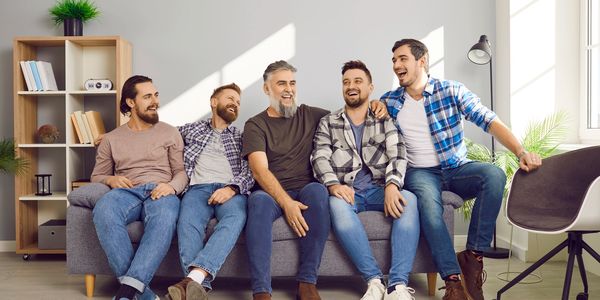 Five men laughing together on a gray couch in a bright room.