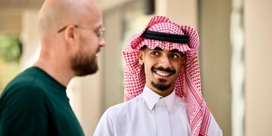 Two men talking outside 1 wearing a green shirt & the other in a red-and-white checkered headscarf 