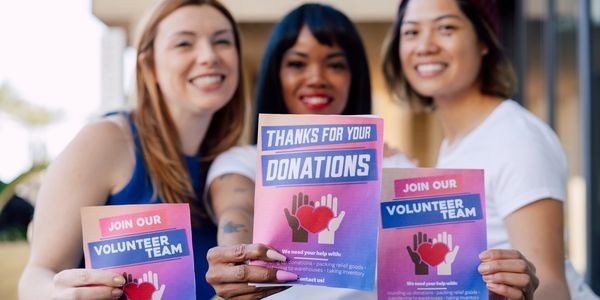 Three smiling women holding volunteer and donation flyers outdoors.