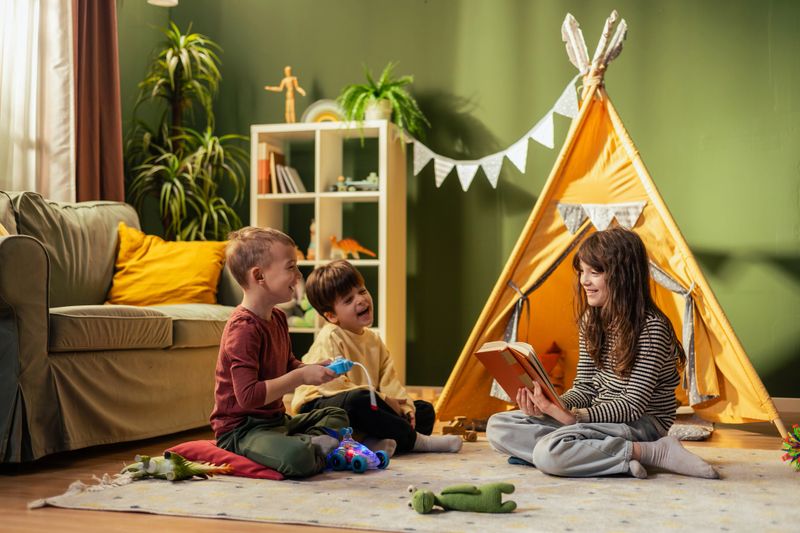 Girl reading a book to two boys playing in a teepee tent, enjoying quality time together in a cozy playroom